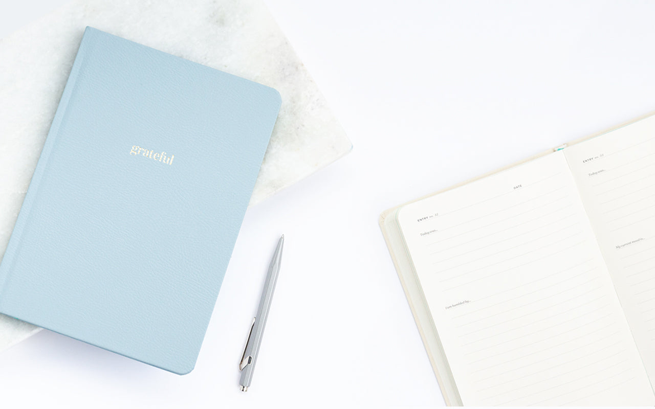 A blue gratitude journal sits on a white desk next to an open Ink+Volt Planner.