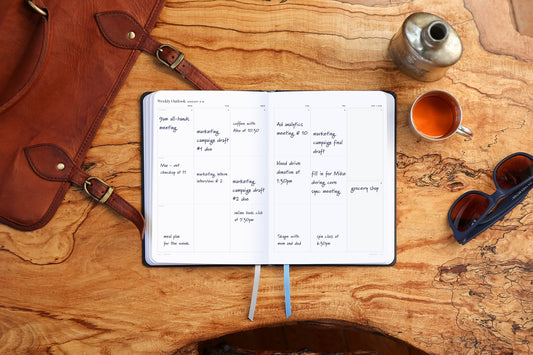 An open planner with many deadlines written in it on a wooden table with a few accessories.