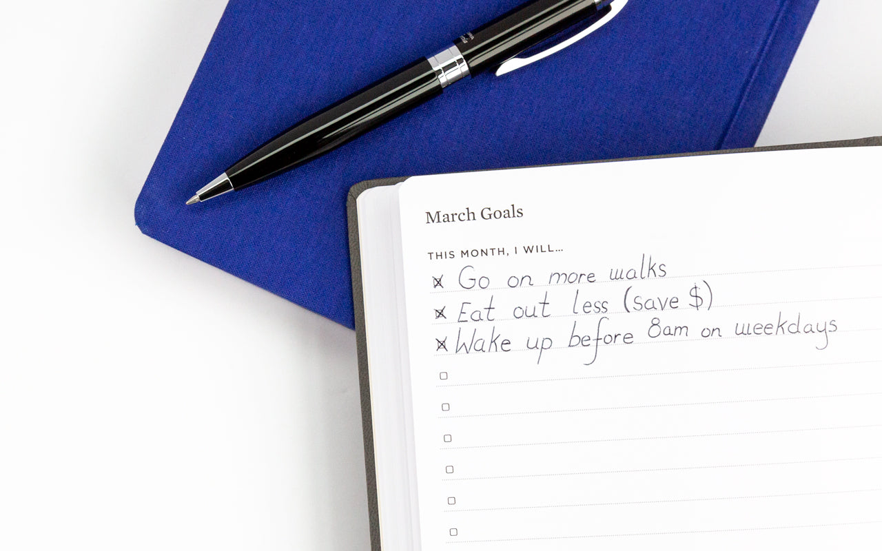 A blue planner and pen sit under an open planner with several goals written on the first page.