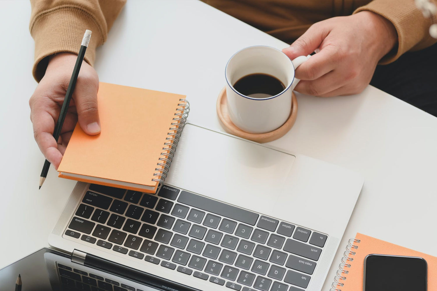 A hand holding a spiral notebook above a laptop with a black pencil and a cup of coffee.