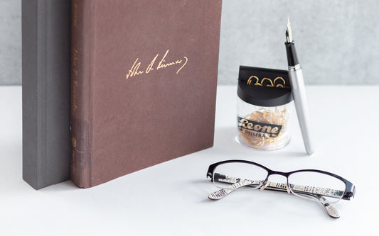 Two old books sit on a desk next to a pair of glasses and a paper clip holder.