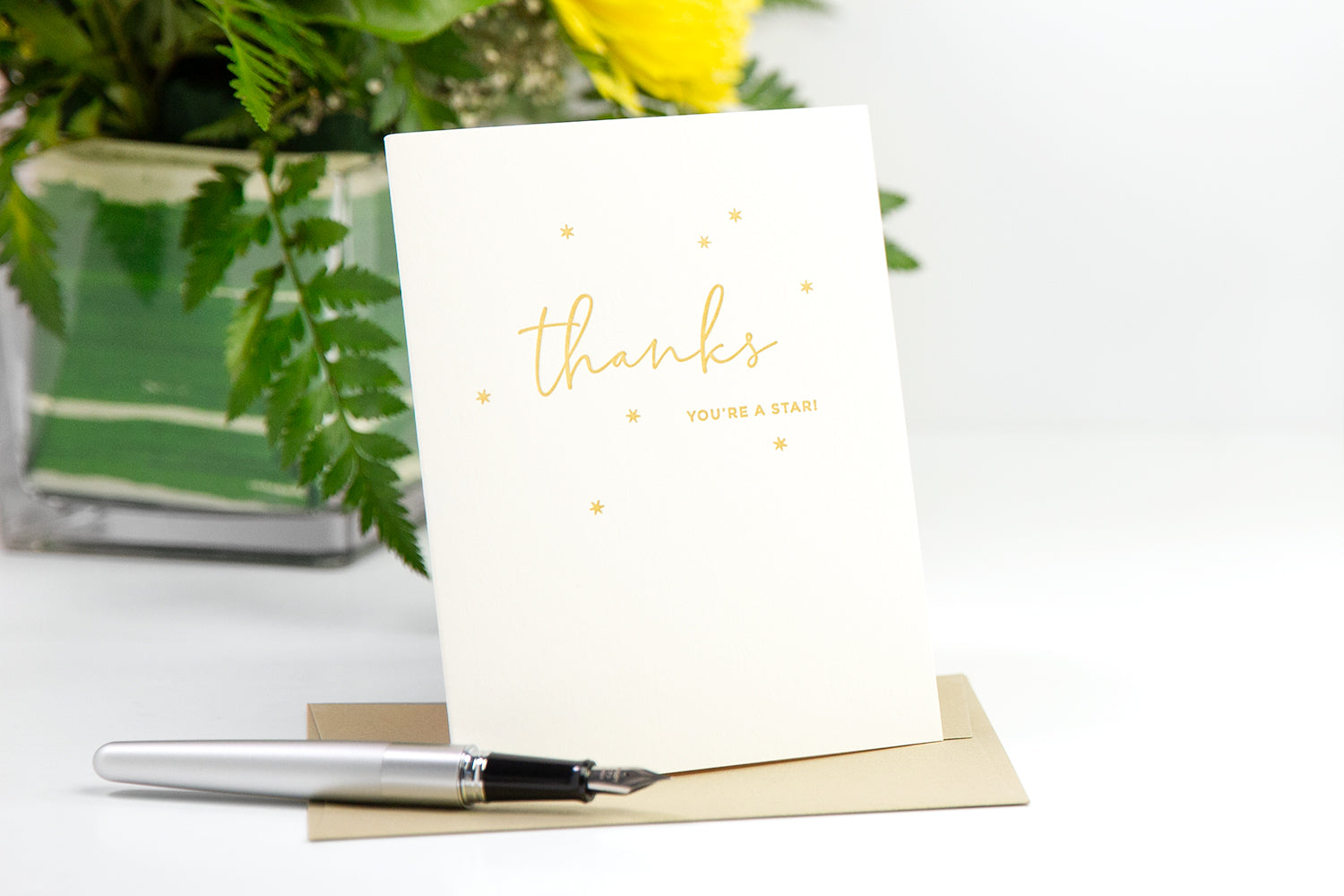 A white thank you card with a gold cursive "thanks" on the front, on a white desk with yellow flowers.
