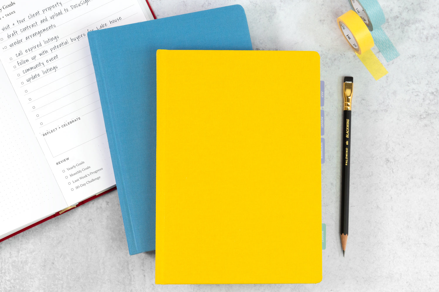 A bright blue planner and a bright yellow planner stacked on a white desk next to a black pencil and to-do list