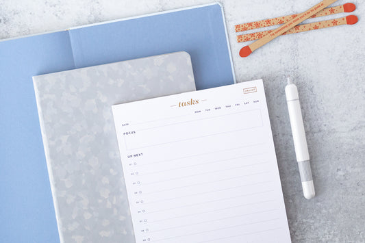 A task pad sits on top of a silver notebook on top of a blue open book, next to a white stylish pen and a "spark courage" matchstick set, all on a white desk.