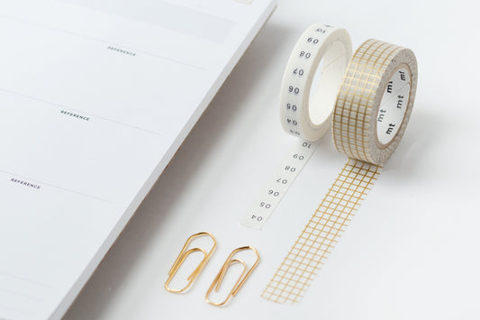 Two rolls of washi tape, rolled out and stuck to a desk next to a calendar and two gold paperclips.