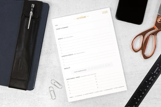 An action notepad on a white desk with an array of other desk accessories including metallic scissors, a black planner, and a pen holder