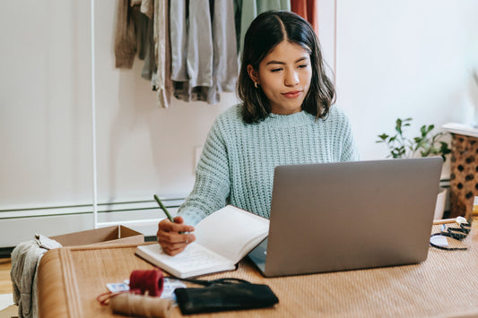 A woman sits at a laptop and writes in a notebook