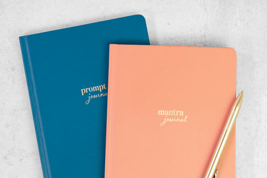 A blue prompt journal and a coral mantra journal on a grey table with a gold pen