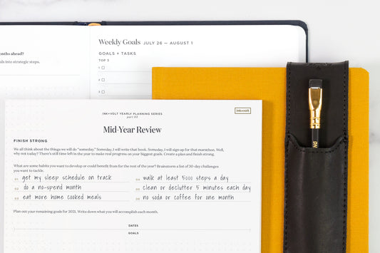 a mid-year review worksheet, partially filled out, on top of a yellow planner with a black pencil holder