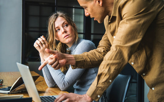 A man and woman talk at a desk as the man points at a laptop screen.