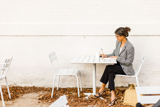 A professionally dressed woman writes in a notebook at a white table outside