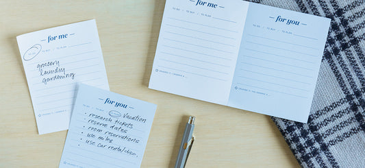 An Ink+Volt For Me For You Task Pad sits on a desk with a checkered cloth. The pages are filled out with handwriting outlining tasks for planning a vacation.