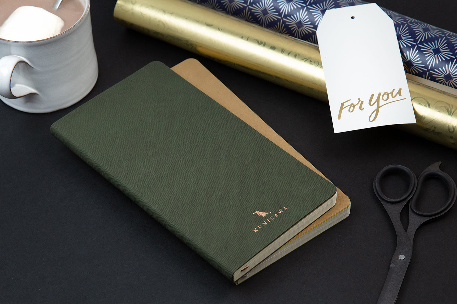 a green kunisawa notebook and a gold kunisawa notebook on a black table with wrapping paper and a gift tag