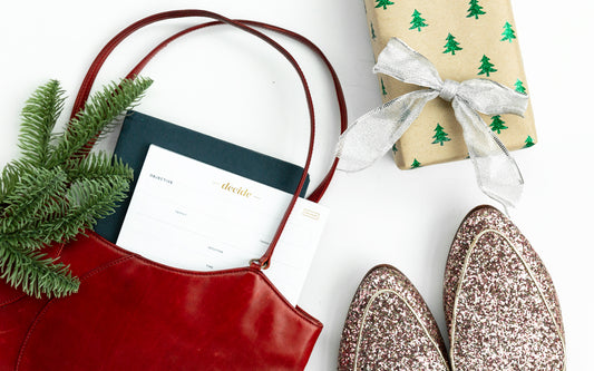 A red bag with a planner and productivity pad, next to a sprig of spruce, glittery shoes, and a gift.