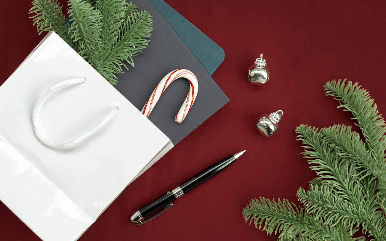 A present in a bag sits on a desk with a pen, jingle bells, and evergreen boughs