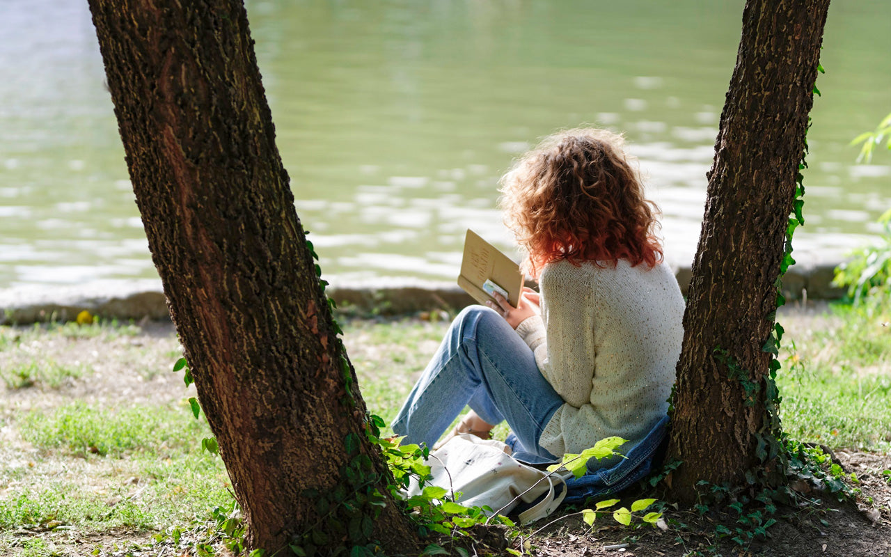 A woman sits by a river reading a book, while leaning up against a tree.