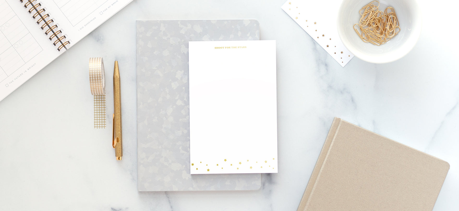 An organized desk with gold accessories, a light grey notebook, and a white and gold notepad.