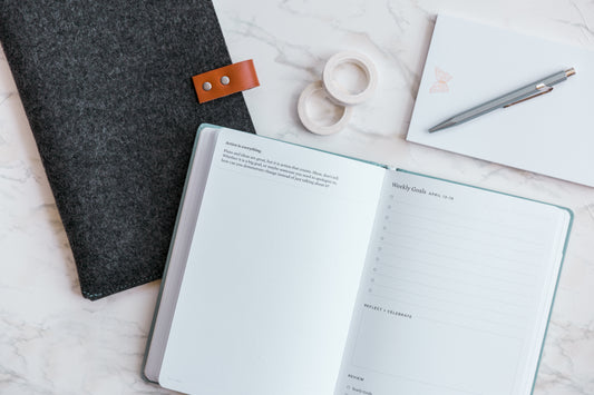 A planner open to the weekly planner pages sits on a marble countertop next to a wool planner sleeve, a pen, and a notepad.