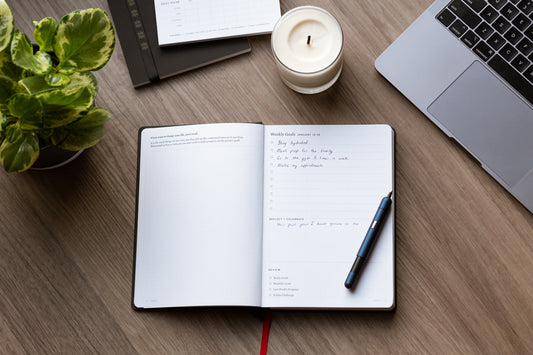 A planner with goals written in it sits on a wood desk next to a green plant, a candle, a laptop, and a blue pen.