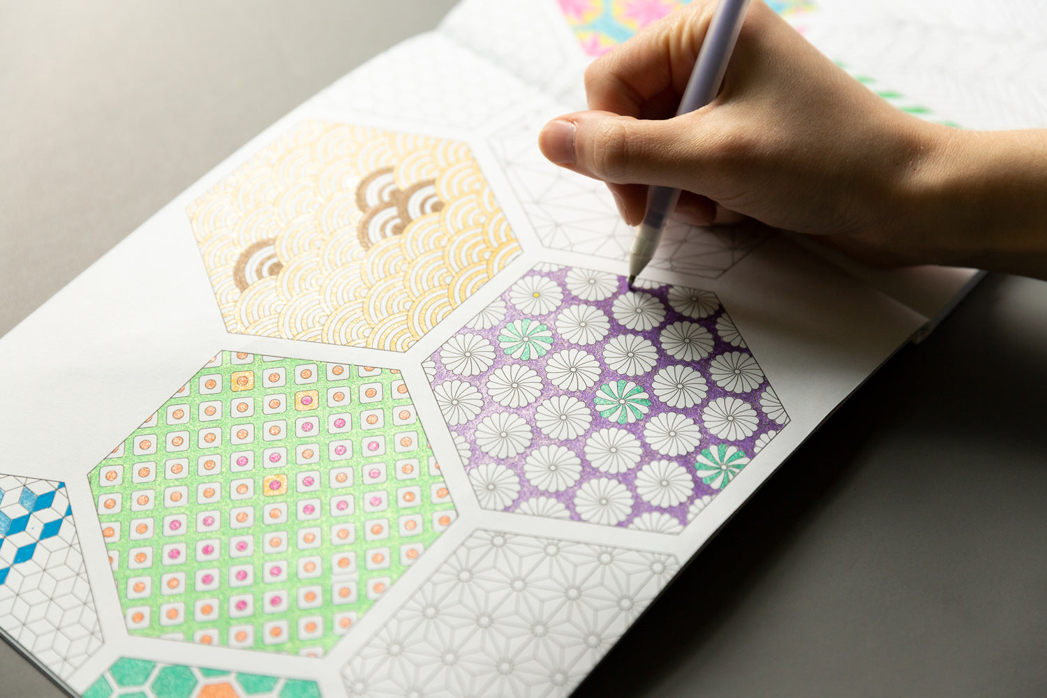 A woman's hand uses a purple colored pencil to color in a pattern of flowers in a coloring book.