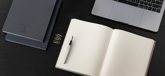 A notebook sits open with a fountain pen on it next to a laptop and two other notebooks.