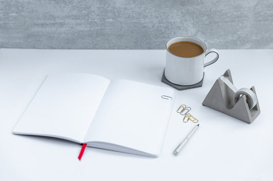 A blank notebook sits open on a desk next to a pen, a cup of coffee, and a modern tape dispenser.