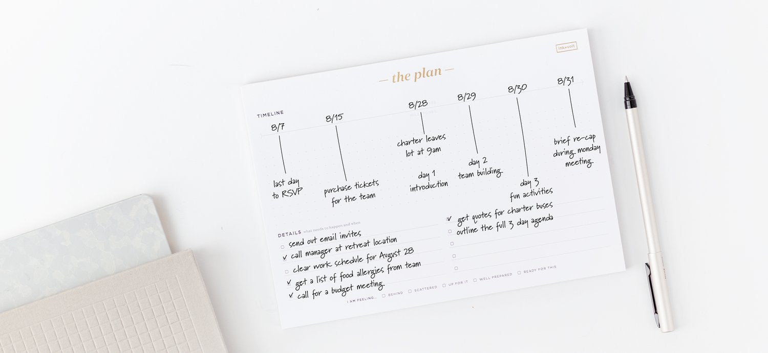 An ink+volt project planing timeline pad sits on a white desk next to a pen, filled out with important project dates.