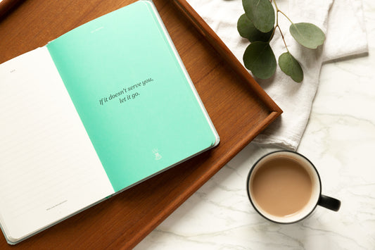 A book sits on a wooden tray next to a cup of tea and a sprig of green. The book is open to a quote about perseverance.