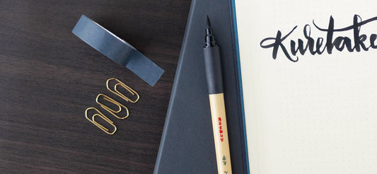 A brush pen on a brown desk with a notebook open to hand-lettered writing