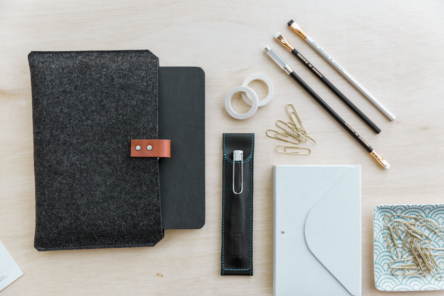 A planner in a wool sleeve, on a wooden desk next to office supplies like pencils, tape, a pen, envelopes, and paper clips.