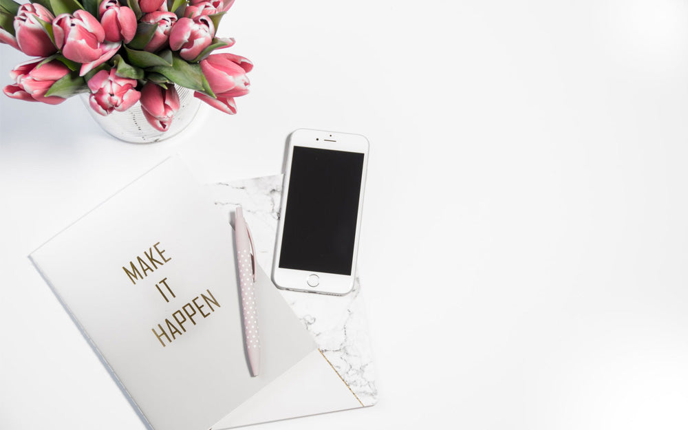A card that says "Make it Happen" lays on a table near tulips, a pen, and a phone