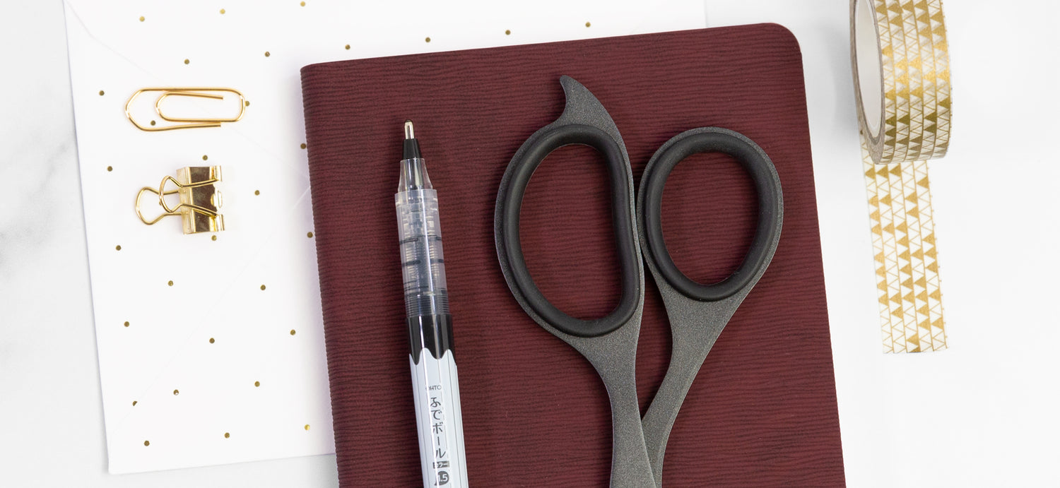 An OHTO rollerball pen sits on a red notebook next to black scissors and gold desk accessories like washi tape.