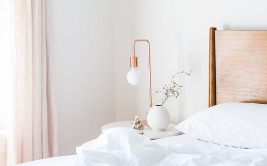 An all-white bedroom, with a bed with white linens and a bedside table with a white vase and rose gold lamp.