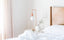 An all-white bedroom, with a bed with white linens and a bedside table with a white vase and rose gold lamp.