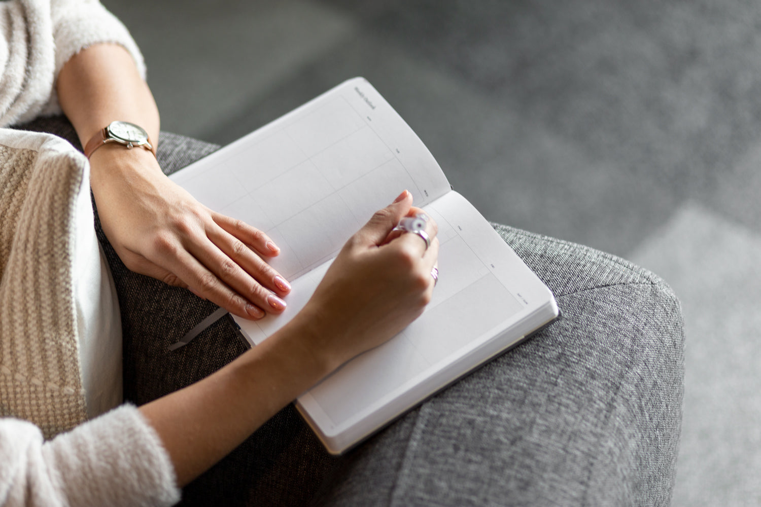 A woman sitting on a grey couch writes in a planner with a pen.