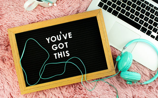 A letterboard with the phrase "You've got this" on it sits on a pink rug next to green headphones and a laptop.