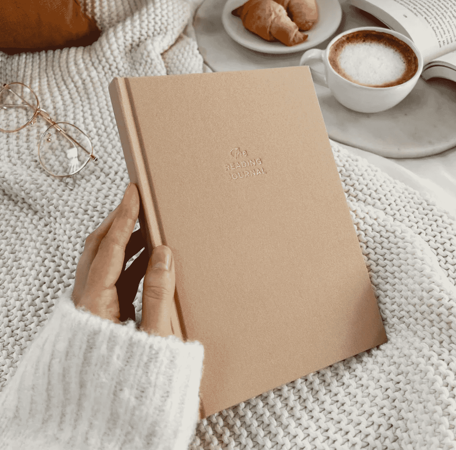 A womans hand holds a guided reading journal on a cozy bed with a cup of coffee