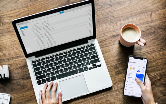 An overhead view of a desk with a laptop showing a full inbox and a hand holding a phone with live data updates, and a cup of coffee.