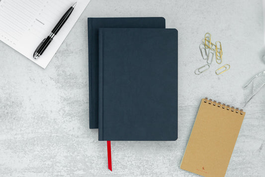 Two navy blue Ink+Volt hardcover notebooks sit on a marble countertop, next to a notepad, planner page, and paper clips.