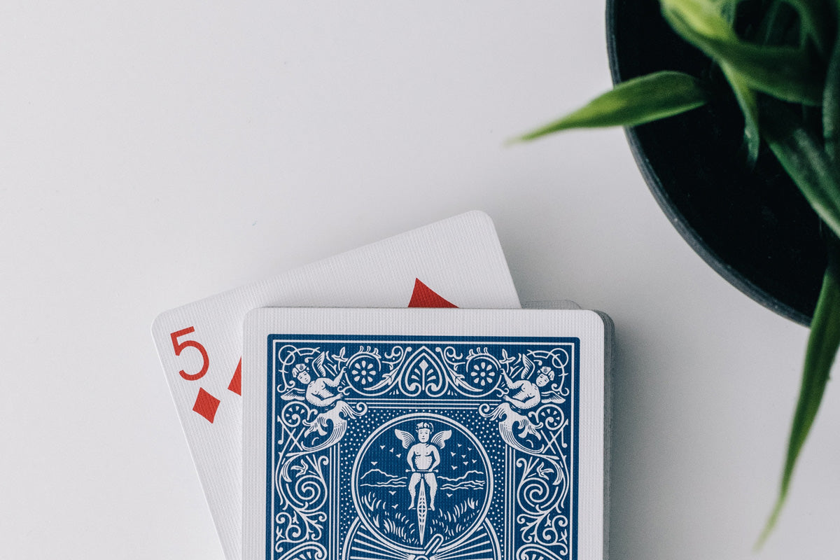 A deck of cards on a white table, with a 5 of diamonds peeking out next to a green plant.