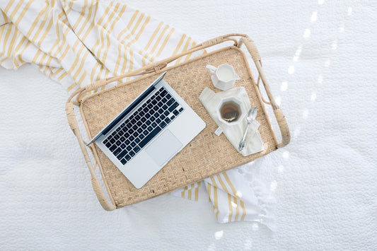 A laptop sits on a wicker tray with a cup of tea on a white sheet.