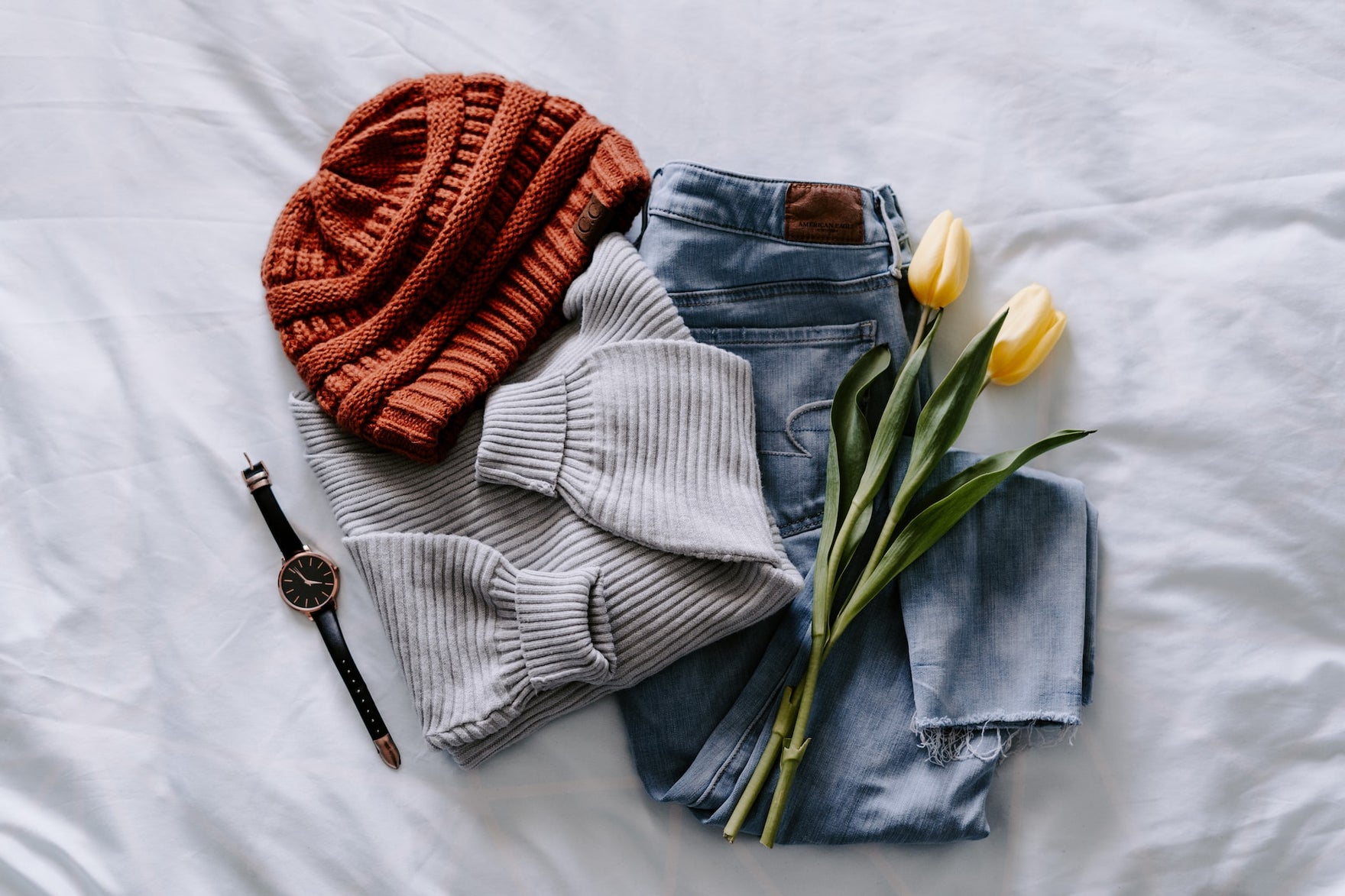 A red hat, grey sweater, jeans, watch, and yellow flowers piled on a white wheet