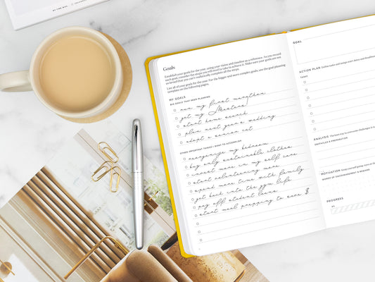 A yellow planner open on a marble tabletop next to a cup of tea and a magazine