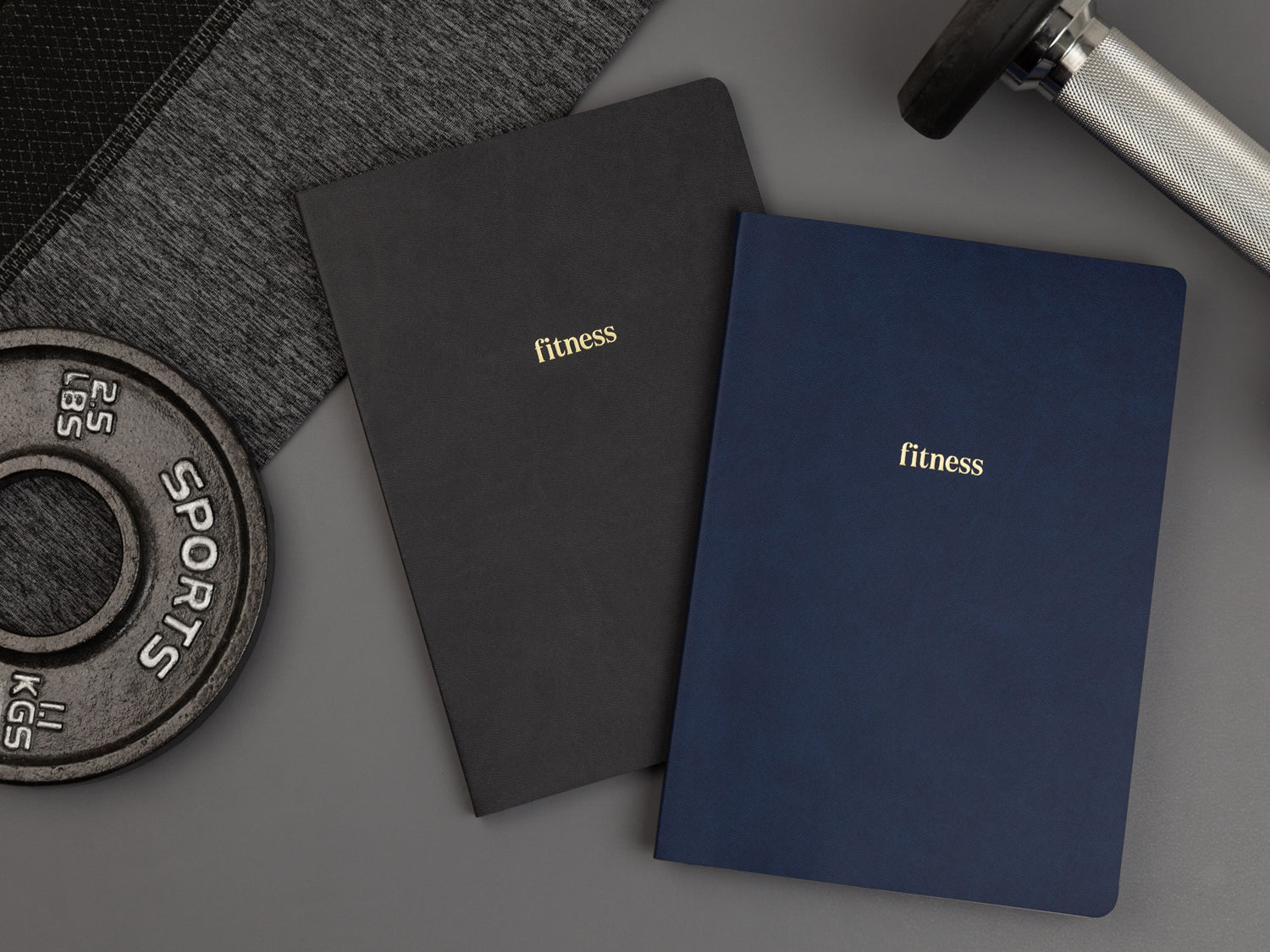 A blue fitness planner and a black fitness planner on a black floor next to two weights