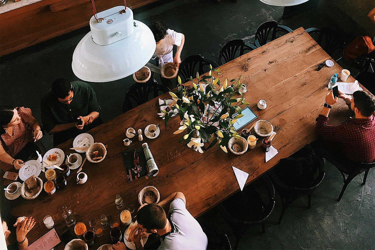 A long wooden table with flowers on it and people working around it