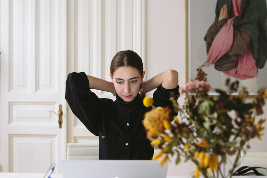 A woman sits at a table with her hands behind her head, looking disappointed