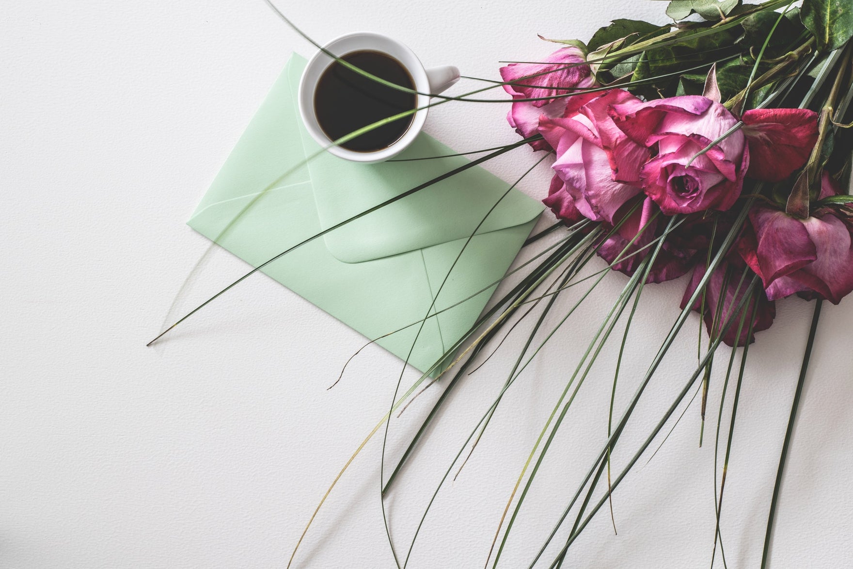 A bouquet of pink roses sits on a table next to a cup of coffee and a sage green envelope.
