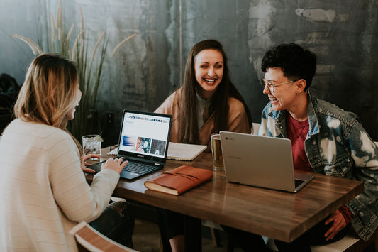 3 people with laptops and drinks sit around a wood table smiling and laughing