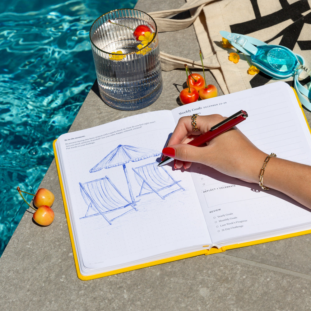 A person sketches in a yellow planner by a swimming pool