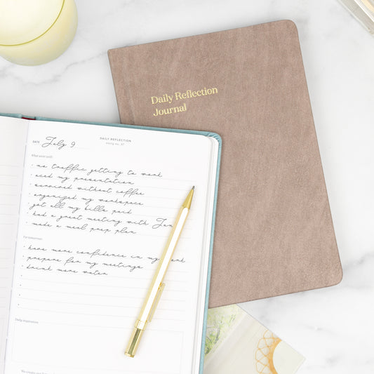 Two daily reflection journals on a white desk, one open to a page with handwritten notes and a gold pen.